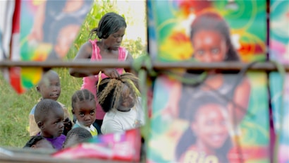A woman is styling another person's hair outdoors, surrounded by several children. The setting is vibrant with colorful posters in the foreground, giving the scene an artistic feel. The background shows greenery, indicating an outdoor rural environment.
