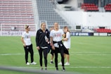 A group of women wearing Washington Spirit apparel are on a soccer field, likely preparing for a game or practice. One woman holds a water bottle and they appear to be in a casual and relaxed mood. There are empty seats in the background, suggesting the setting is a stadium.