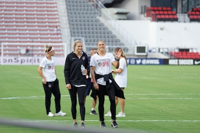 A group of women wearing Washington Spirit apparel are on a soccer field, likely preparing for a game or practice. One woman holds a water bottle and they appear to be in a casual and relaxed mood. There are empty seats in the background, suggesting the setting is a stadium.