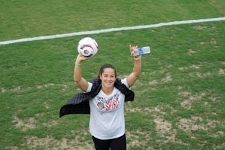 A person on a soccer field is holding a soccer ball in one hand and a water bottle in the other. The person is wearing a white shirt with 'Washington Spirit' written on it and is draped with a jacket over the shoulders. They are smiling towards the camera.