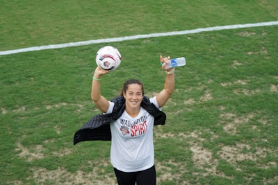 A person on a soccer field is holding a soccer ball in one hand and a water bottle in the other. The person is wearing a white shirt with 'Washington Spirit' written on it and is draped with a jacket over the shoulders. They are smiling towards the camera.