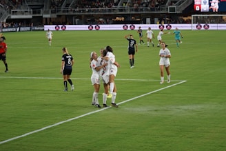 A group of female soccer players in white uniforms are celebrating on the field, suggesting they just scored a goal. Other players in black uniforms are walking around, and a referee in red is also present. The match is taking place in a stadium with visible advertising boards and spectators in the stands.