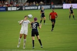 Two soccer players are engaged in a match on a grassy field, with one player attempting to head the ball while an opponent positions herself nearby. A referee in a red shirt observes the game along with other players in the background. Advertising banners and a crowd of spectators are visible along the sidelines.
