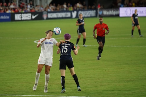Two soccer players are engaged in a match on a grassy field, with one player attempting to head the ball while an opponent positions herself nearby. A referee in a red shirt observes the game along with other players in the background. Advertising banners and a crowd of spectators are visible along the sidelines.
