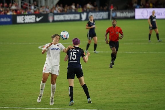 Two soccer players are engaged in a match on a grassy field, with one player attempting to head the ball while an opponent positions herself nearby. A referee in a red shirt observes the game along with other players in the background. Advertising banners and a crowd of spectators are visible along the sidelines.