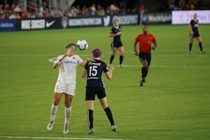 Two soccer players are engaged in a match on a grassy field, with one player attempting to head the ball while an opponent positions herself nearby. A referee in a red shirt observes the game along with other players in the background. Advertising banners and a crowd of spectators are visible along the sidelines.