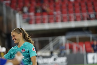 A smiling woman in a turquoise sports jersey with a ponytail on a sports field. The background shows a stadium with red seats and some blurred spectators.
