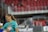 A smiling woman in a turquoise sports jersey with a ponytail on a sports field. The background shows a stadium with red seats and some blurred spectators.