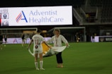 A soccer player wearing a white jersey with the number 12 and the name 'Sullivan' holds a large, unfurled banner on a sports field. Another person stands opposite, helping with the banner. The stadium is partially empty, with a large advertising screen in the background displaying 'Adventist HealthCare'.