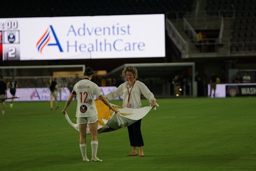 A soccer player wearing a white jersey with the number 12 and the name 'Sullivan' holds a large, unfurled banner on a sports field. Another person stands opposite, helping with the banner. The stadium is partially empty, with a large advertising screen in the background displaying 'Adventist HealthCare'.