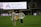 A soccer player wearing a white jersey with the number 12 and the name 'Sullivan' holds a large, unfurled banner on a sports field. Another person stands opposite, helping with the banner. The stadium is partially empty, with a large advertising screen in the background displaying 'Adventist HealthCare'.