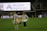 A soccer player wearing a white jersey with the number 12 and the name 'Sullivan' holds a large, unfurled banner on a sports field. Another person stands opposite, helping with the banner. The stadium is partially empty, with a large advertising screen in the background displaying 'Adventist HealthCare'.