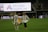 A soccer player wearing a white jersey with the number 12 and the name 'Sullivan' holds a large, unfurled banner on a sports field. Another person stands opposite, helping with the banner. The stadium is partially empty, with a large advertising screen in the background displaying 'Adventist HealthCare'.