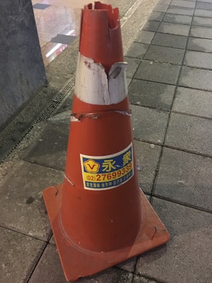 A damaged orange traffic cone is placed on a paved sidewalk. The top part of the cone is broken and there are visible scratches and dirt marks on it. A label with text and contact information is attached to the cone.