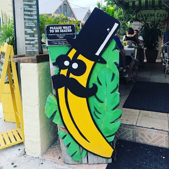A whimsical sign in the shape of a banana character with a top hat and mustache is prominently displayed near the entrance of a caf&eacute;. The sign is colorful, with the banana being bright yellow and surrounded by green leaves. Behind the sign is a table with people sitting and enjoying their meals in an outdoor seating area surrounded by plants.