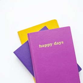 Several colorful notebooks are stacked on a white background. The top notebook is bright pink with the words 'happy days' embossed in gold on the cover. Beneath it, a purple notebook is visible, followed by a yellow one at the bottom.