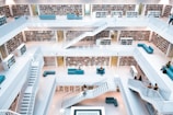 A modern multi-level library with white walls and railings. Bookshelves filled with books line the walls on each floor. There are several teal seating areas scattered throughout, and people are seated or walking up the stairs. The design is open with a central atrium providing a clear view of each level.