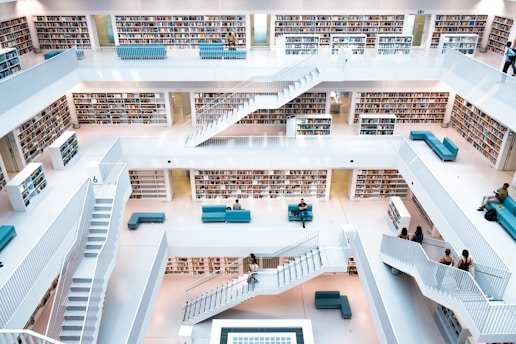 A modern multi-level library with white walls and railings. Bookshelves filled with books line the walls on each floor. There are several teal seating areas scattered throughout, and people are seated or walking up the stairs. The design is open with a central atrium providing a clear view of each level.