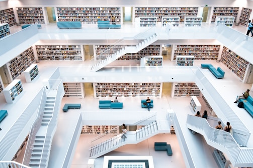 A modern multi-level library with white walls and railings. Bookshelves filled with books line the walls on each floor. There are several teal seating areas scattered throughout, and people are seated or walking up the stairs. The design is open with a central atrium providing a clear view of each level.