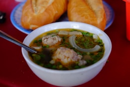 A bowl of soup with meatballs, green onions, and slices of onion is placed on a red surface. Two pieces of baguette are on a plate in the background. A spoon is resting in the bowl, indicating it is ready to eat.