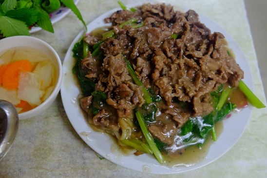 A plate filled with a stir-fried dish featuring thinly sliced beef and vibrant green vegetables in a savory sauce. To the side is a bowl containing pickled vegetables with visible slices of carrot and other various colored vegetables. Fresh herbs are partially visible in the background.