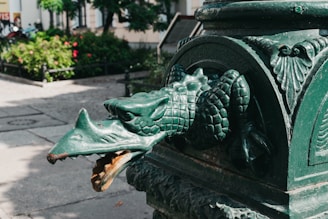 A detailed statue of a dragon's head protrudes from a concrete base in an outdoor urban setting. The dragon appears to be made of metal, painted green, and exhibits intricate scales and fierce facial features. The background shows part of a garden with green plants and flowers, as well as some trees, and pathways. The environment is sunlit, with shadows cast on the ground.