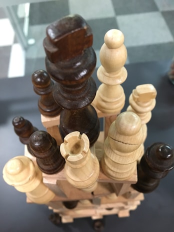 Wooden chess pieces are stacked in a tower formation, with both light and dark pieces arranged in an intricate manner. The chessboard in the background has a checkerboard pattern with alternating light and dark squares.