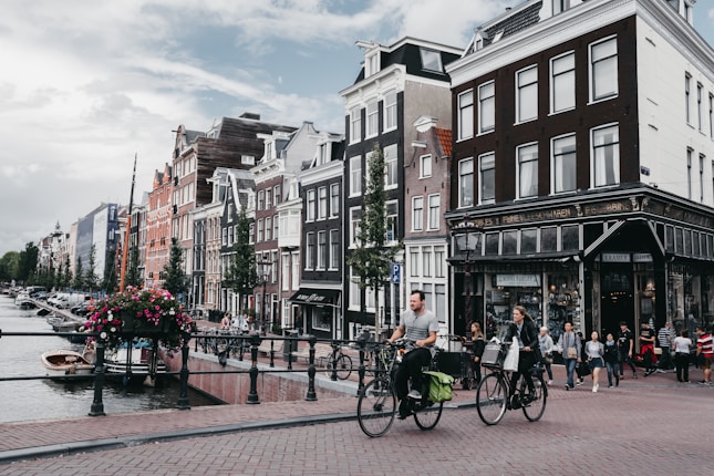 Cyclists ride along a canal lined with traditional Dutch buildings, featuring large windows and gabled roofs. The street is bustling with pedestrians, and flowers hang over the railing of the canal. The sky is overcast, adding a serene quality to the scene.