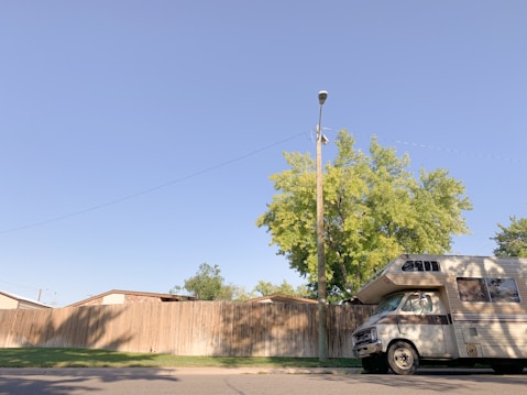 A motorhome is parked on a street beside a high wooden fence. A wooden utility pole stands nearby, and a tree with green leaves is visible behind the fence. The sky is clear and blue.