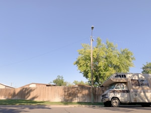 A motorhome is parked on a street beside a high wooden fence. A wooden utility pole stands nearby, and a tree with green leaves is visible behind the fence. The sky is clear and blue.