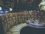 A futuristic interior featuring a curved booth with a padded design, reminiscent of a spacecraft or sci-fi setting. A table with illuminated buttons and lights is at the center. The walls contain various panels, pipes, and glowing elements, enhancing the technological aesthetic.