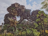 A fantastical landscape featuring floating rocky islands covered in lush vegetation. Vines and exotic plants drape from the rocks, and a whimsical tree-like plant with colorful flowers stands prominently in the foreground. The bright sky with scattered clouds adds to the surreal atmosphere.