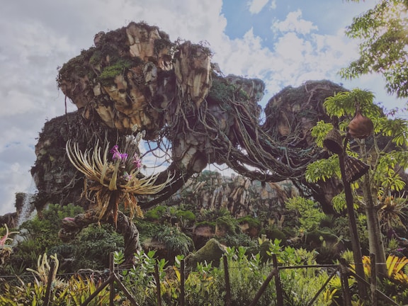 A fantastical landscape featuring floating rocky islands covered in lush vegetation. Vines and exotic plants drape from the rocks, and a whimsical tree-like plant with colorful flowers stands prominently in the foreground. The bright sky with scattered clouds adds to the surreal atmosphere.