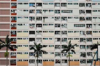 A high-rise residential building with multiple balconies, each having different colors and items such as air conditioning units and clothes. The facade consists of repeating rectangular patterns. Several palm trees are in the foreground, adding greenery to the otherwise urban scene.