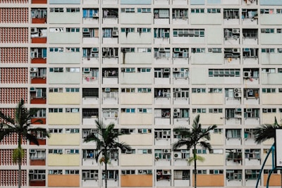 A high-rise residential building with multiple balconies, each having different colors and items such as air conditioning units and clothes. The facade consists of repeating rectangular patterns. Several palm trees are in the foreground, adding greenery to the otherwise urban scene.