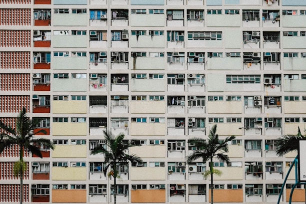 A high-rise residential building with multiple balconies, each having different colors and items such as air conditioning units and clothes. The facade consists of repeating rectangular patterns. Several palm trees are in the foreground, adding greenery to the otherwise urban scene.