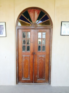 A rustic wooden door with intricate carved panels and a semi-circular stained glass window above it. The glass window features a mix of colors, including red, blue, and green sections. The door is set in a plain, light-colored wall, flanked by two framed pictures on either side.