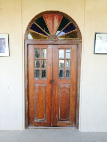 A rustic wooden door with intricate carved panels and a semi-circular stained glass window above it. The glass window features a mix of colors, including red, blue, and green sections. The door is set in a plain, light-colored wall, flanked by two framed pictures on either side.