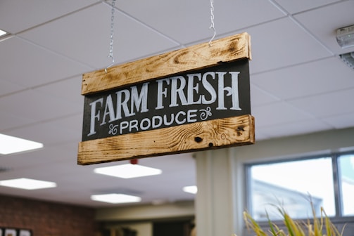 A wooden sign with black background and white text that reads 'FARM FRESH PRODUCE' is suspended from the ceiling with chains. The sign has a rustic appearance, with visible wood grain and metal fittings. The setting appears to be indoors with ceiling lights and a window in the background.