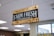 A wooden sign with black background and white text that reads 'FARM FRESH PRODUCE' is suspended from the ceiling with chains. The sign has a rustic appearance, with visible wood grain and metal fittings. The setting appears to be indoors with ceiling lights and a window in the background.