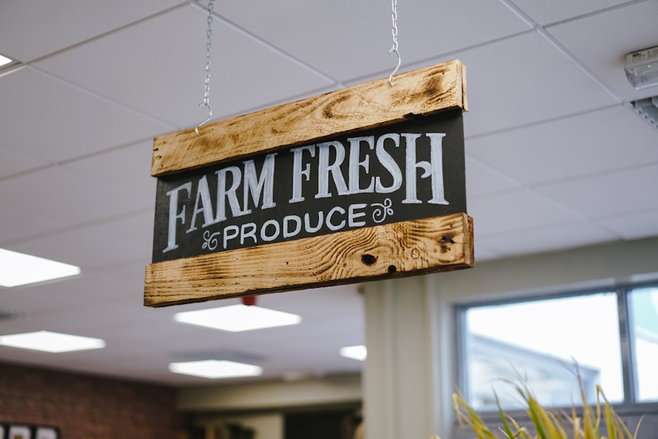 A wooden sign with black background and white text that reads 'FARM FRESH PRODUCE' is suspended from the ceiling with chains. The sign has a rustic appearance, with visible wood grain and metal fittings. The setting appears to be indoors with ceiling lights and a window in the background.