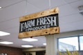 A wooden sign with black background and white text that reads 'FARM FRESH PRODUCE' is suspended from the ceiling with chains. The sign has a rustic appearance, with visible wood grain and metal fittings. The setting appears to be indoors with ceiling lights and a window in the background.