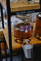 A large glass carboy filled with a golden liquid is placed on a wooden shelf, with a metal spigot attached. Below the spigot hangs a small metal bucket. The liquid appears to be some kind of oil or beverage and is labeled with a purple sticker that reads 'Yare Valley Oils'. The setting is rustic, with other bottles in the background.