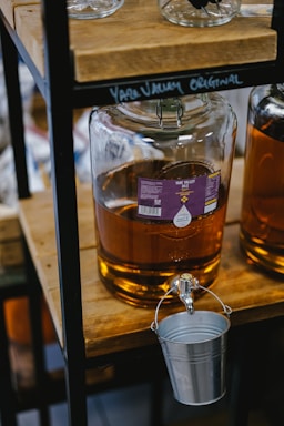 A large glass carboy filled with a golden liquid is placed on a wooden shelf, with a metal spigot attached. Below the spigot hangs a small metal bucket. The liquid appears to be some kind of oil or beverage and is labeled with a purple sticker that reads 'Yare Valley Oils'. The setting is rustic, with other bottles in the background.