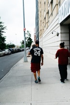 A man carrying a small dog walks on a sidewalk next to a tall building with a parking sign. He is wearing a dark t-shirt with the name 'Sanchez' and the number '24' on it, along with red shorts and black shoes. Next to him is another person wearing a red top. Parked cars line the street, and there are trees in the background.