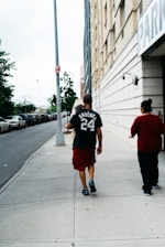 A man carrying a small dog walks on a sidewalk next to a tall building with a parking sign. He is wearing a dark t-shirt with the name 'Sanchez' and the number '24' on it, along with red shorts and black shoes. Next to him is another person wearing a red top. Parked cars line the street, and there are trees in the background.