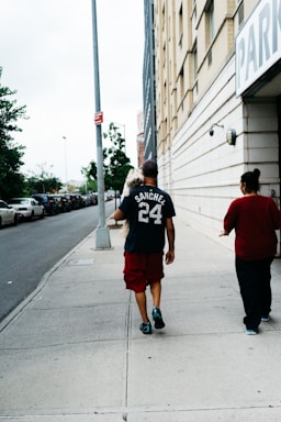 A man carrying a small dog walks on a sidewalk next to a tall building with a parking sign. He is wearing a dark t-shirt with the name 'Sanchez' and the number '24' on it, along with red shorts and black shoes. Next to him is another person wearing a red top. Parked cars line the street, and there are trees in the background.