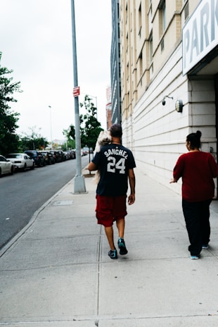 A man carrying a small dog walks on a sidewalk next to a tall building with a parking sign. He is wearing a dark t-shirt with the name 'Sanchez' and the number '24' on it, along with red shorts and black shoes. Next to him is another person wearing a red top. Parked cars line the street, and there are trees in the background.