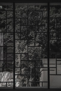 A geometric grid pattern window frame creates a detailed silhouette against a backdrop of lush, textured trees. The scene includes an industrial structure and brick wall on each side, with some tarps or coverings at the bottom, partially visible through the grid.