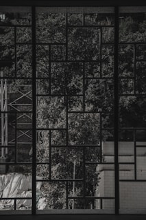 A geometric grid pattern window frame creates a detailed silhouette against a backdrop of lush, textured trees. The scene includes an industrial structure and brick wall on each side, with some tarps or coverings at the bottom, partially visible through the grid.